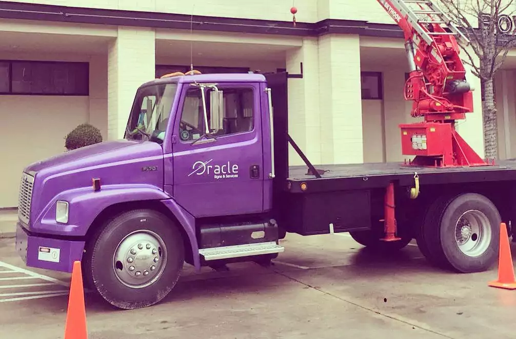 Oracle Signs Texas - Fabrication / Installation of a sign on the side of building. The picture shows a purple construction ruck with a red crane on the bed of the truck.